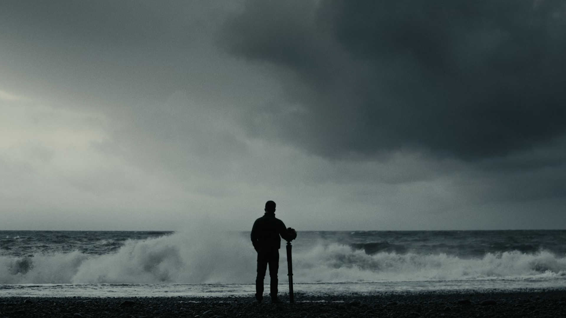 A silhouette of a man with a tripod-mounted camera standing in front of a dark and stormy sea.