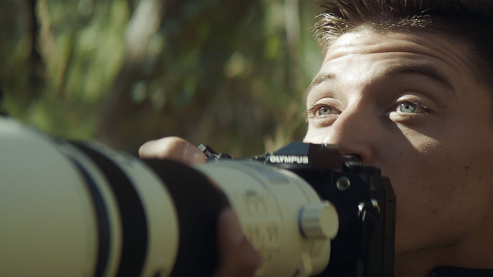 A man with blue eyes looks over his Olympus camera and telephoto lens.