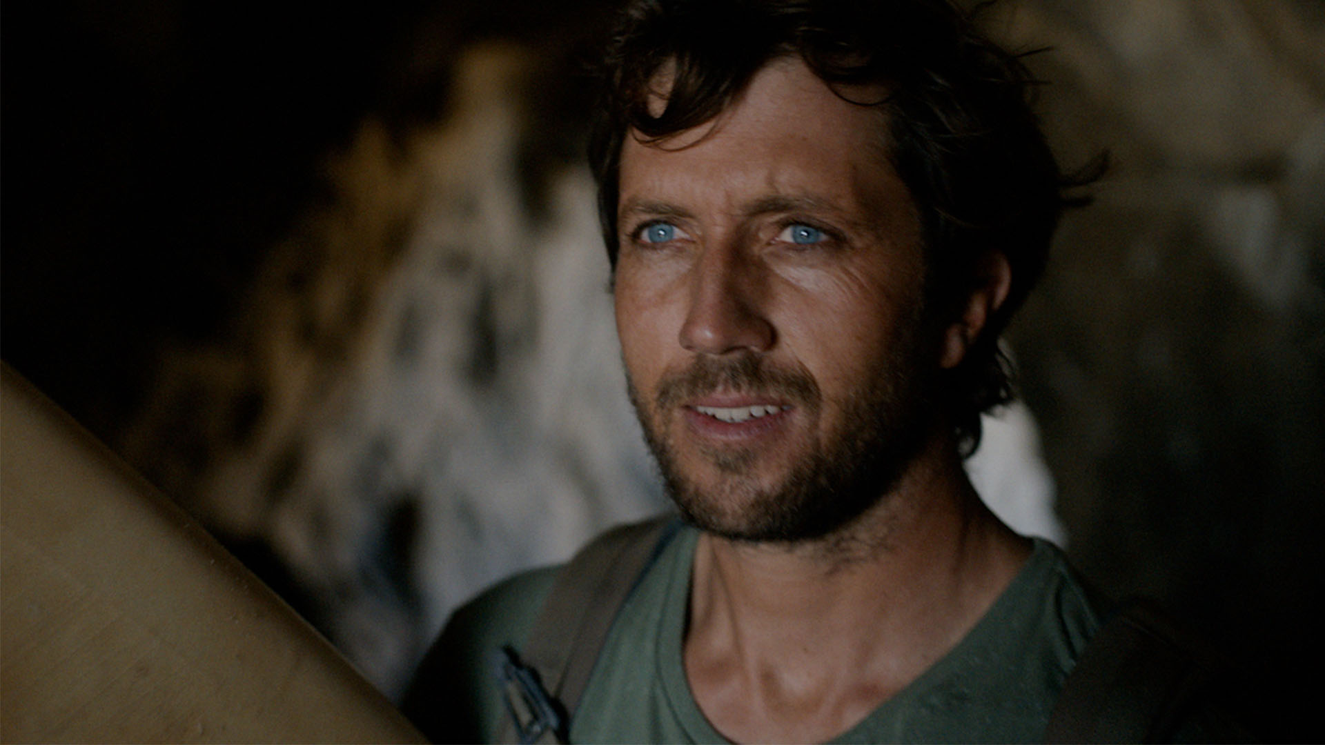 A bearded man with blue eyes looking into the distance inside a cave.