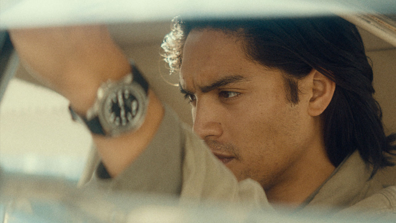 A man with long hair raises his arm with a watch up as he drives through the desert.