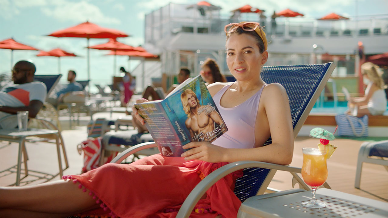 A woman in a beach chair on a cruise ship looks at the camera, holding a romance novel.