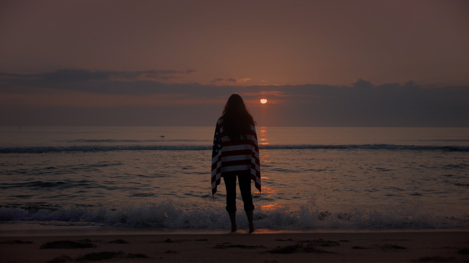 A woman looks out over the horizon at a sunset, a U.S.A. flag draped over her shoulders.