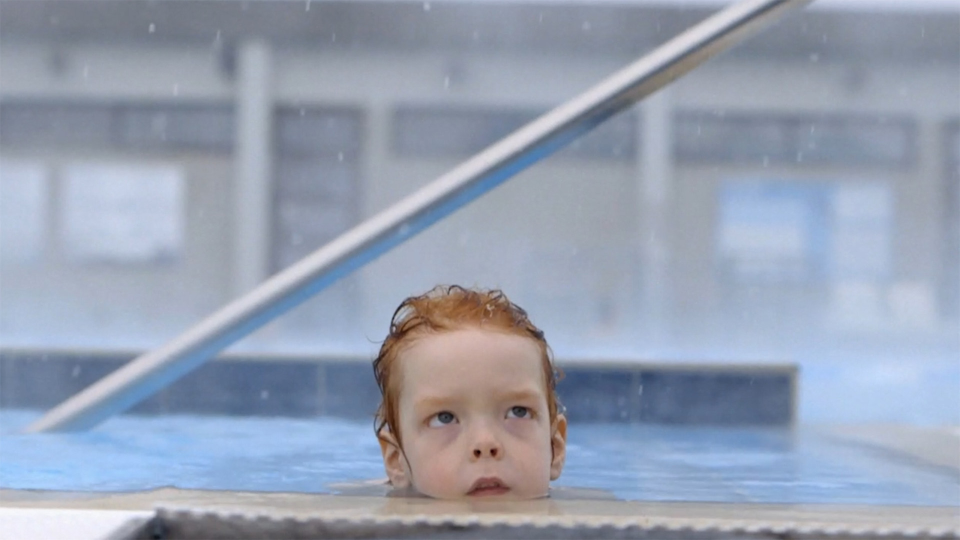 A young boy with orange hair sticks his head out of a heated pool as snow falls.