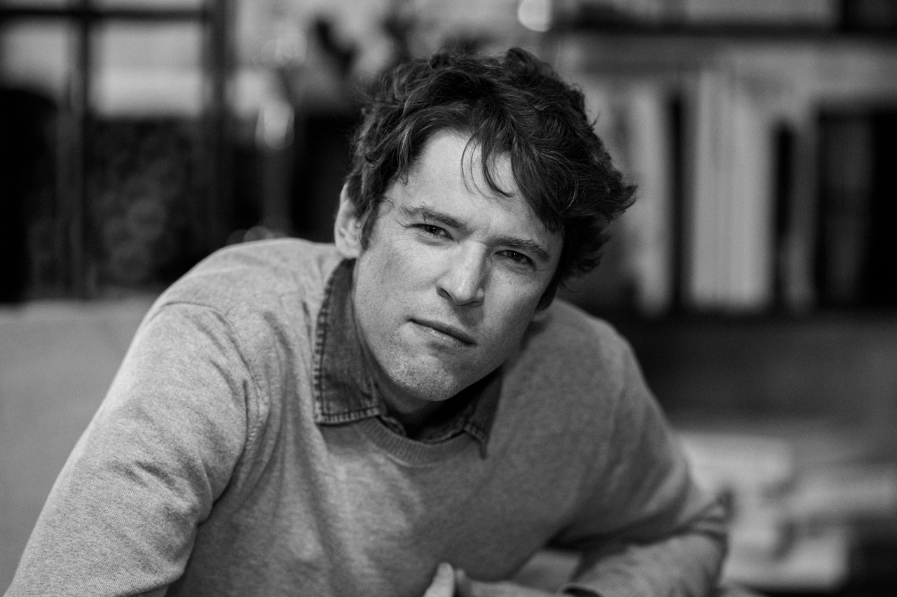 A man with wavy hair wearing a sweater over a collared shirt sits indoors, leaning slightly forward and looking directly at the camera with a thoughtful expression. The image is in black and white.