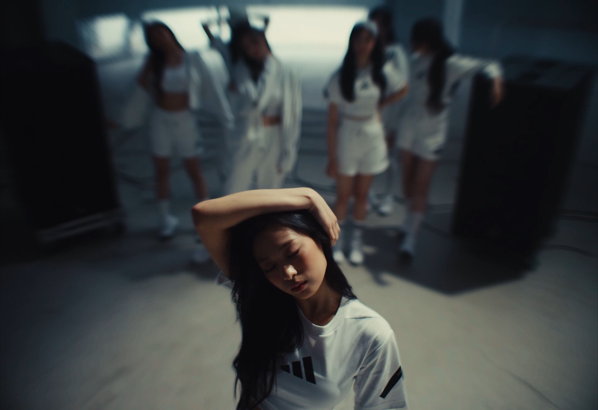 A woman in a white sports outfit stands in the foreground with her eyes closed and one arm over her head, while four other women dressed in white are blurred in the background. The scene has a soft, moody atmosphere.