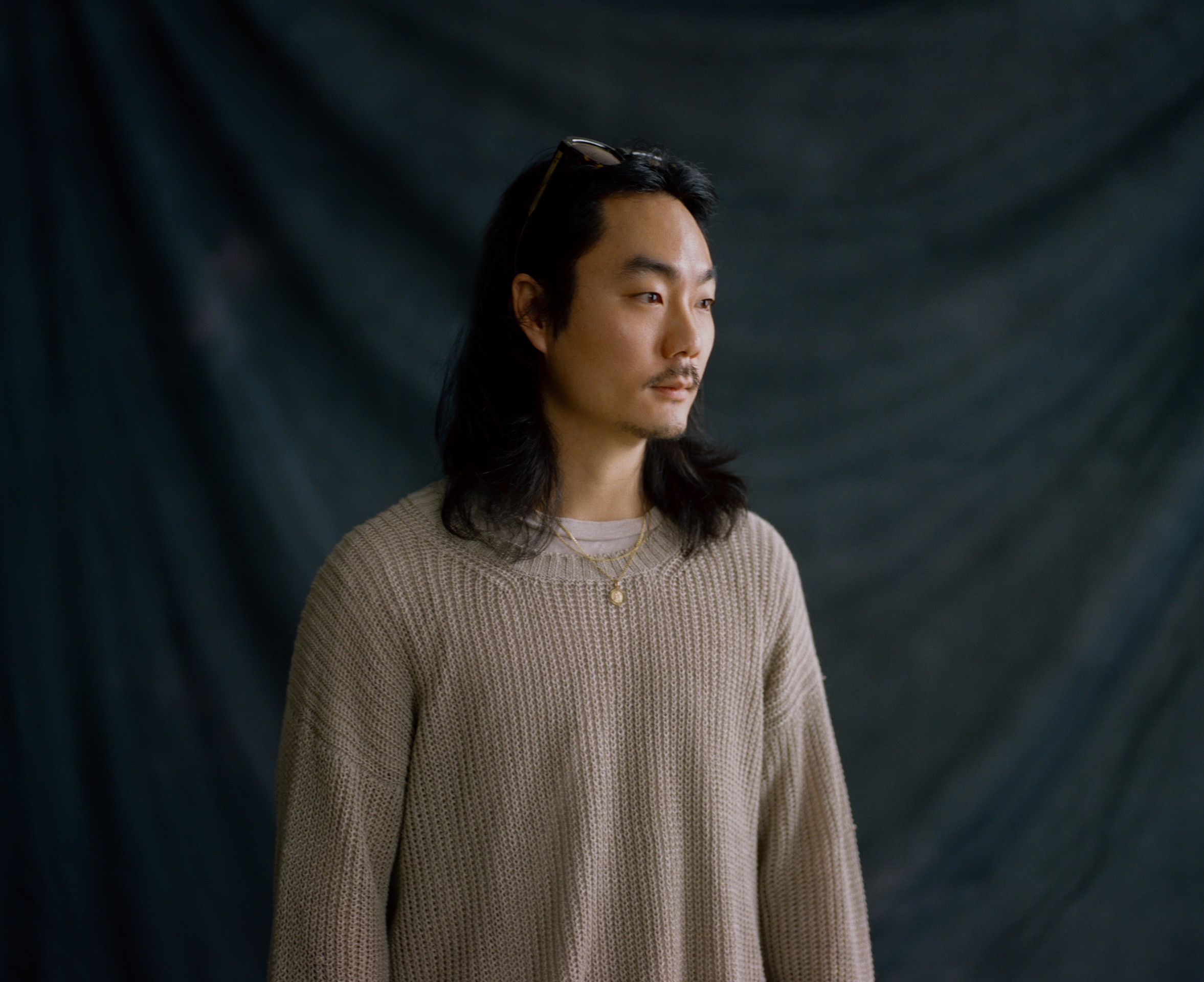 A person with long dark hair, wearing a beige knit sweater and a necklace, stands in front of a dark, textured backdrop, looking slightly to the side with a neutral expression.