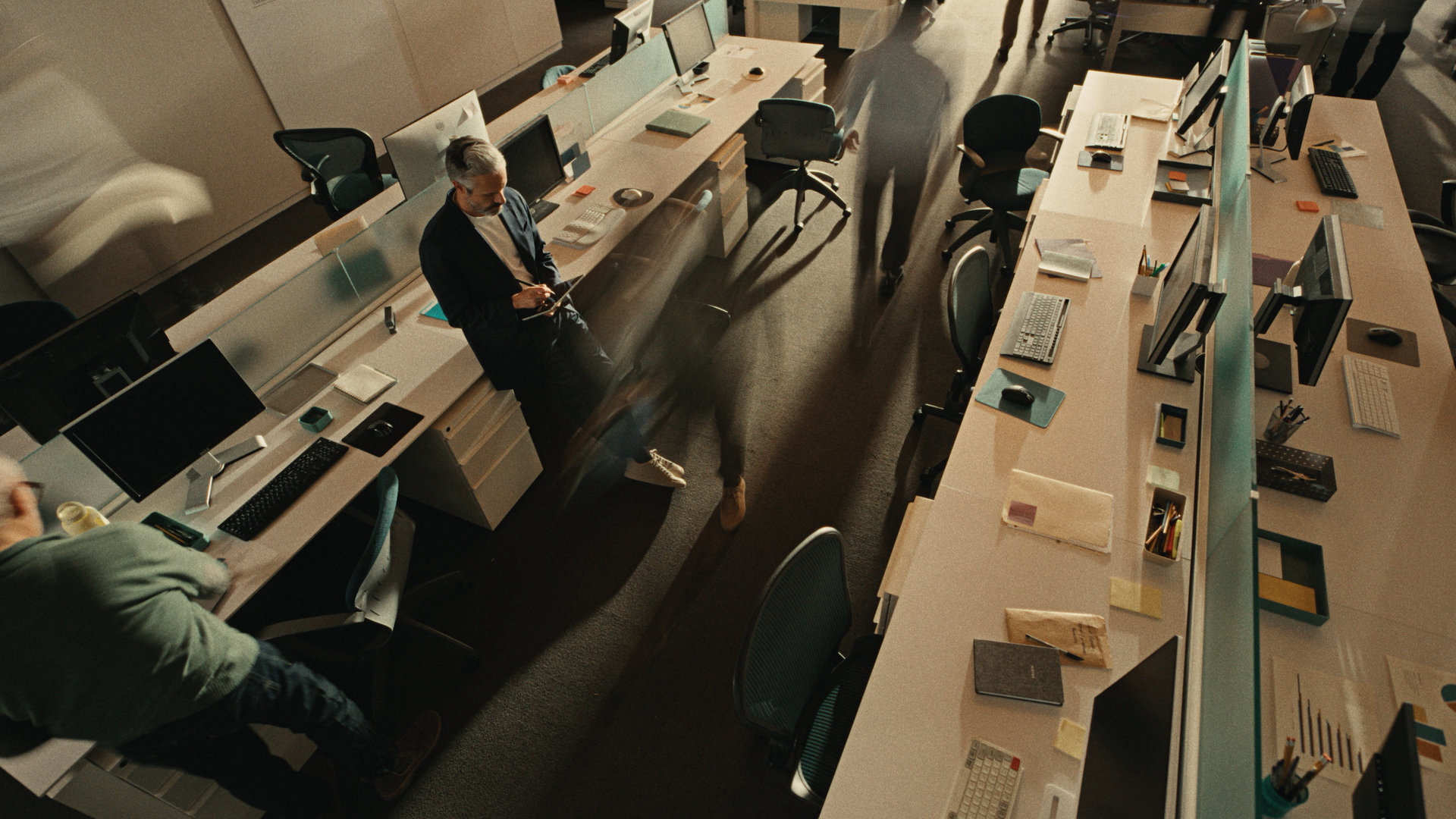 A man stands looking at his phone in a modern, mostly empty office with long desks, computers, chairs, and scattered office supplies as blurred figures walk past, suggesting movement and activity.