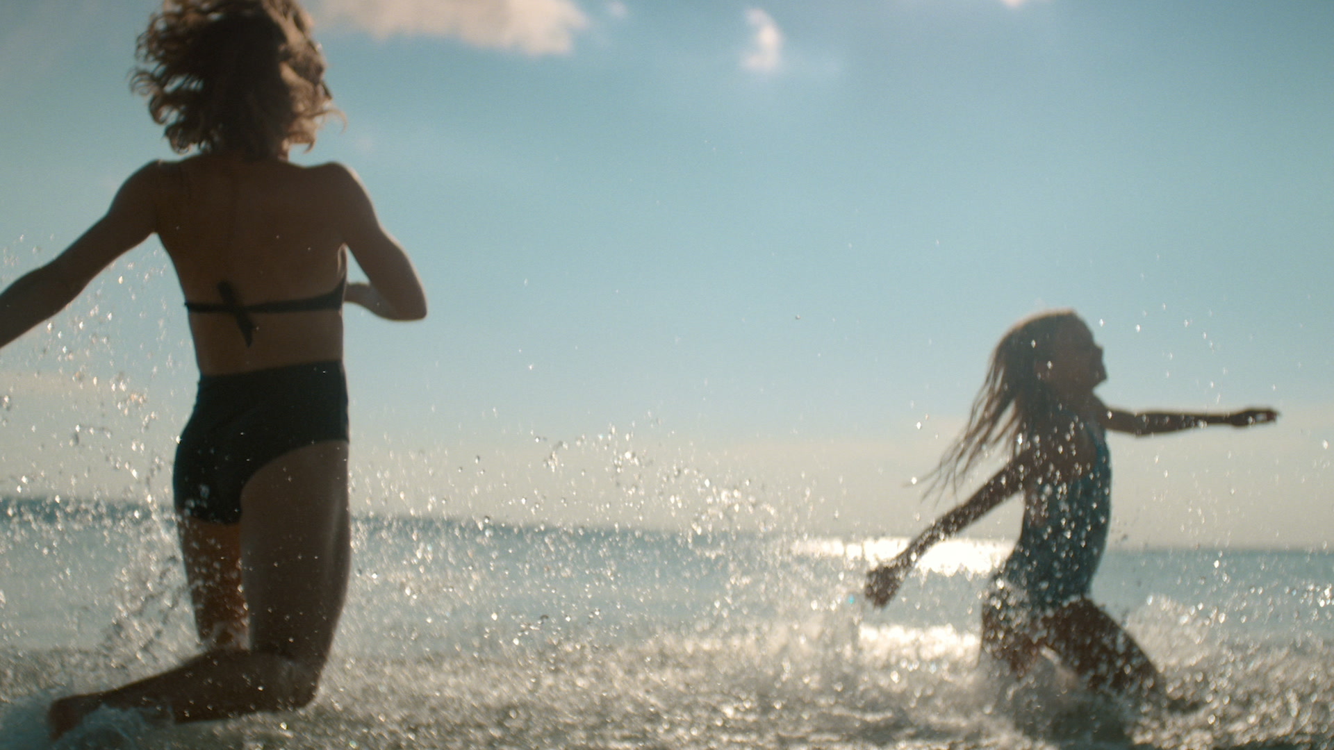 Two children in swimsuits play and splash in the shallow water at the beach on a sunny day, with sunlight reflecting off the sea in the background.