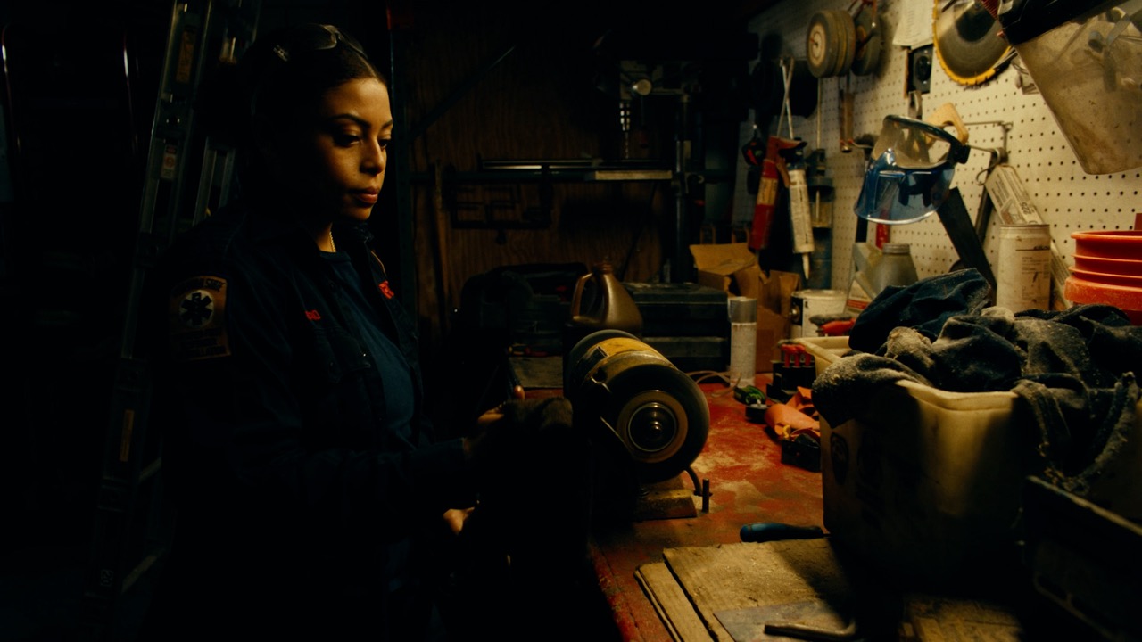 A woman in a dimly lit workshop stands near a workbench with tools and equipment, holding a cloth beside a bench grinder.