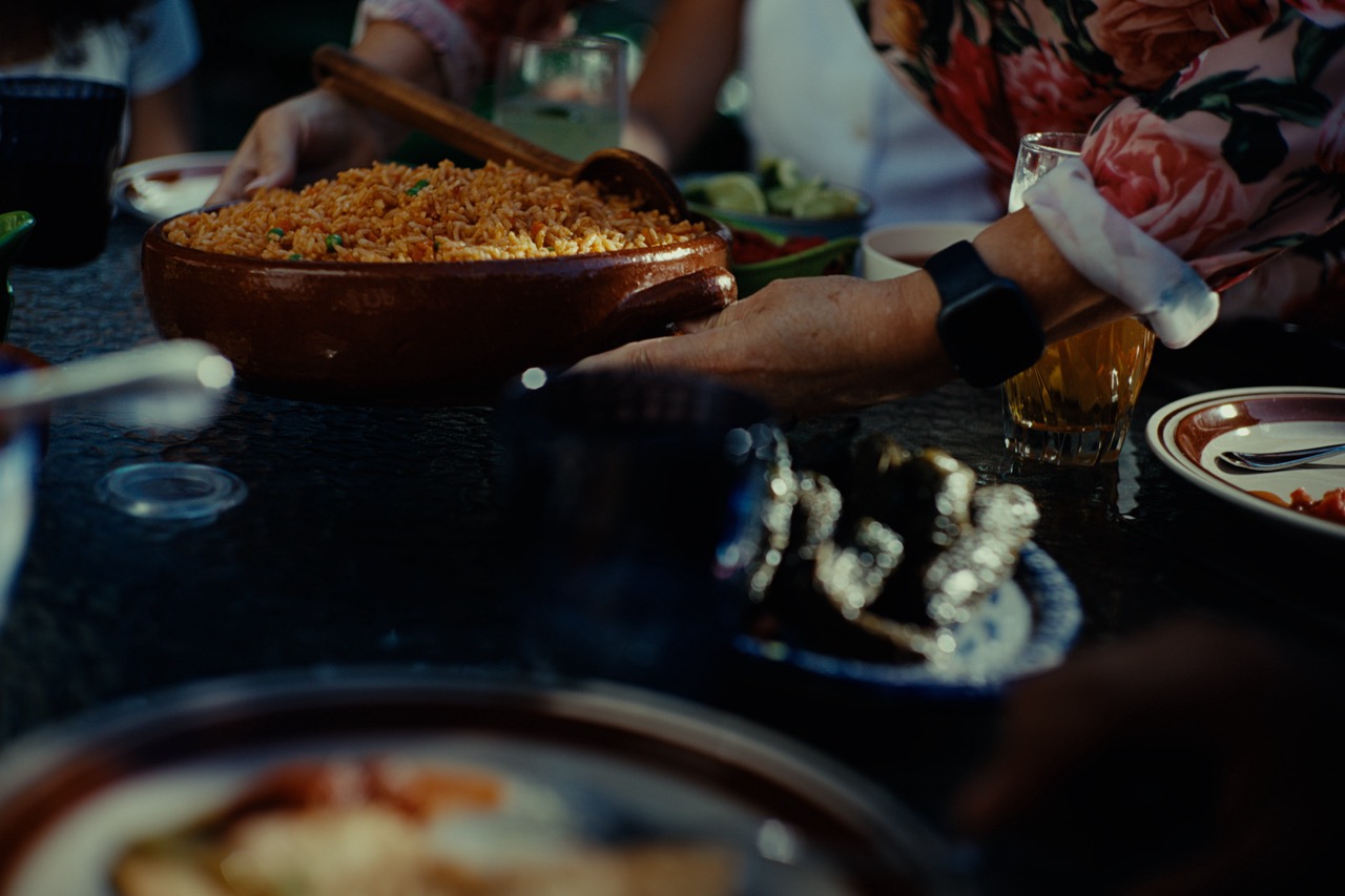A person in a floral shirt serves rice from a ceramic dish at a table set with plates, drinks, and food, suggesting a shared meal or gathering.