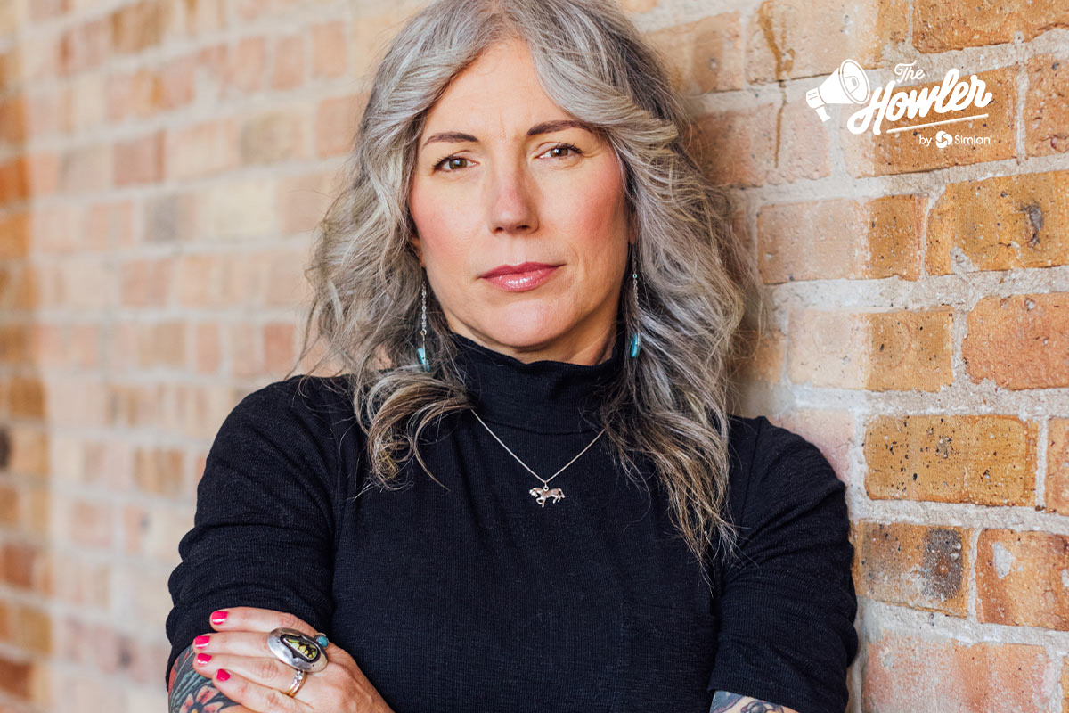 A woman with long gray hair in a black turtleneck stands with arms crossed against a brick wall. She wears jewelry and looks confidently at the camera. “The Howler” logo appears in the top right corner.