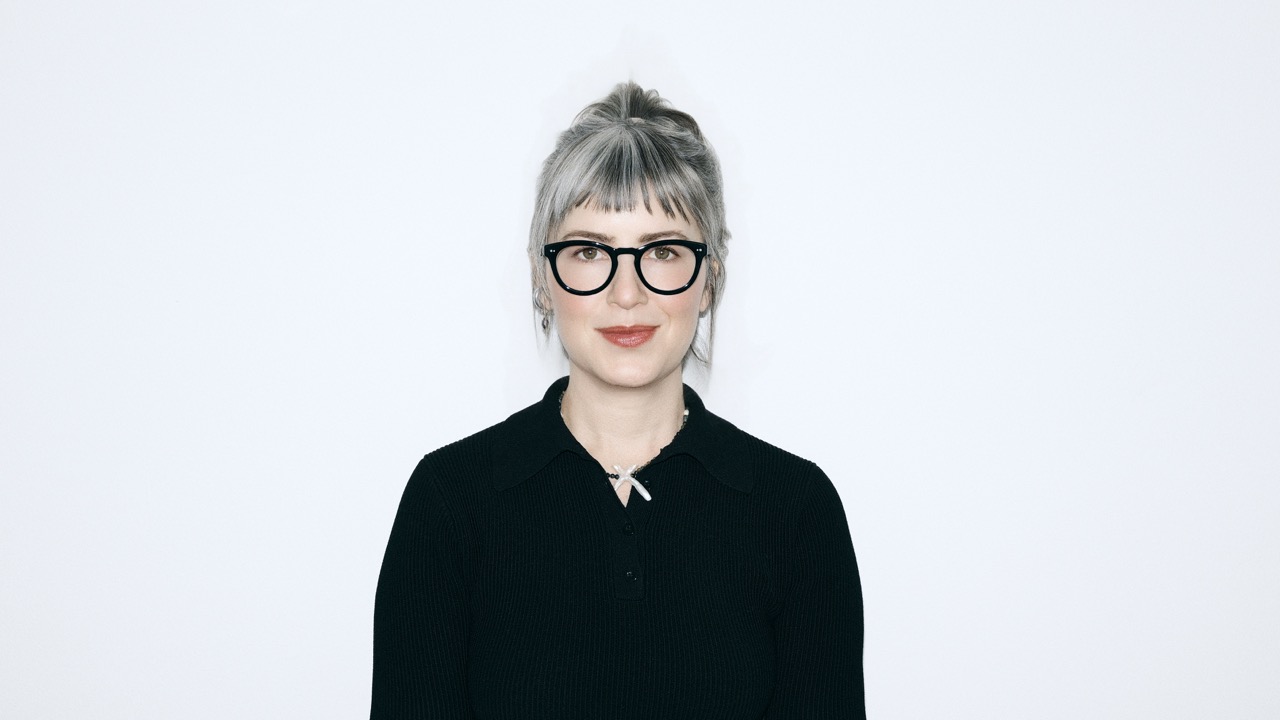 A person with gray hair and black-rimmed glasses smiling against a white background.